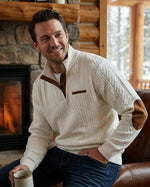 Man sitting by a fireplace wearing a white sweater with brown accents