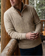 Man in a beige sweater holding a mug on a wooden deck with a fireplace and snowy trees in the background